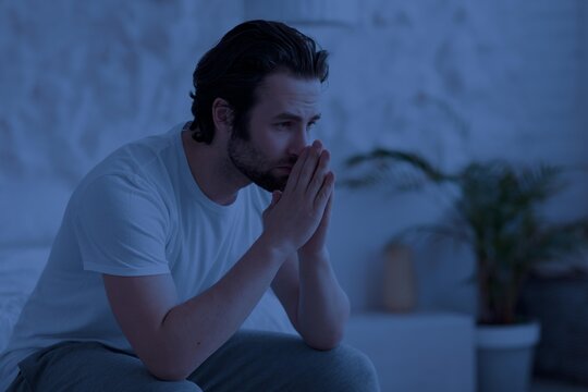Worried Young Caucasian Man Sitting On Bed At Nighttime, Holding Hands Together And Looking Thoughtful, Deep In Thought In Bedroom. Mental Wellness And Stress Relief Concept.