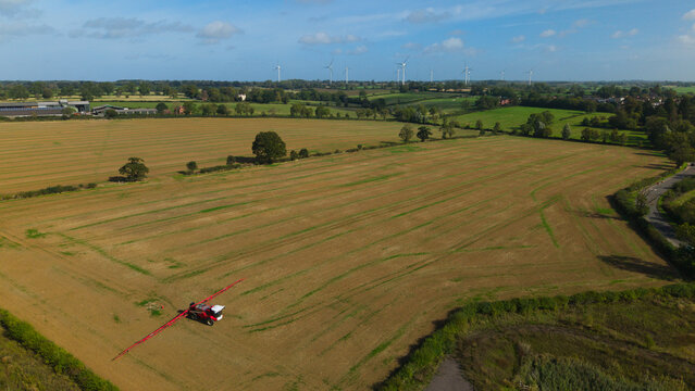 Aerial view of a self propelled agricultural sprayer working in a large harvested field with wind turbines in the distance and green hedgerows in Lutterworth, England, United Kingdom.