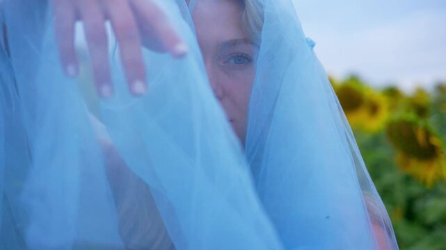 Soft blue veil shrouds a woman's face. Her hand gently brushes the fabric. Sunflowers bloom behind her in golden rows. Her gaze is calm, serene, and thoughtful. Ideal for dreamy, romantic