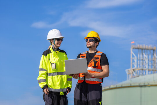 Engineering team in hard hats and safety vests using laptop computer at gas refinery plant, Male technician holding laptop and discussing site inspection with female colleague outdoors