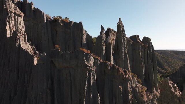 Close aerial movement reveals sunlit earth pillars and deep shadowed clefts at Putangirua Pinnacles, New Zealand. Sparse shrubs cling to ledges above a wooded valley.