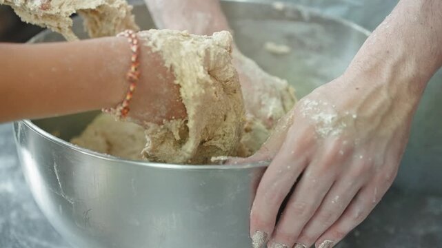 Close view of a woman's hands with long manicured nails and a beaded bracelet working a soft dough in a stainless steel bowl on a floured counter.