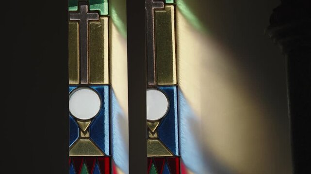Two narrow stained glass windows showing a cross, a white communion host and a gold chalice in blue and yellow panes, light falling on a dark church wall.