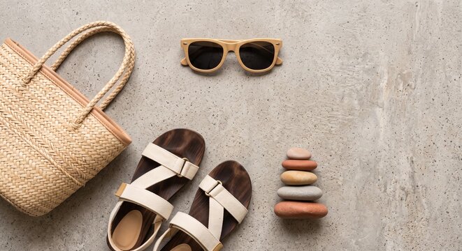 A flat lay of summer essentials includes a woven bag, stylish sunglasses, and comfortable sandals on a concrete surface. This arrangement evokes a sense of relaxation and beach readiness.