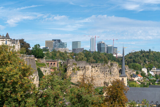 Panoramic view of Luxembourg City, Luxembourg, featuring historic stone fortifications, dramatic cliffs and a church spire above the Grund valley. Modern office towers and construction cranes rise bey