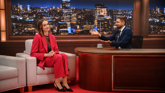 Late Night Talk Show Interview: White Male Host Presenter Gestures Toward Female Celebrity Guest Smiling in the Armchair During a Fun Chat, Glowing City Skyline Backdrop on Television Studio Set