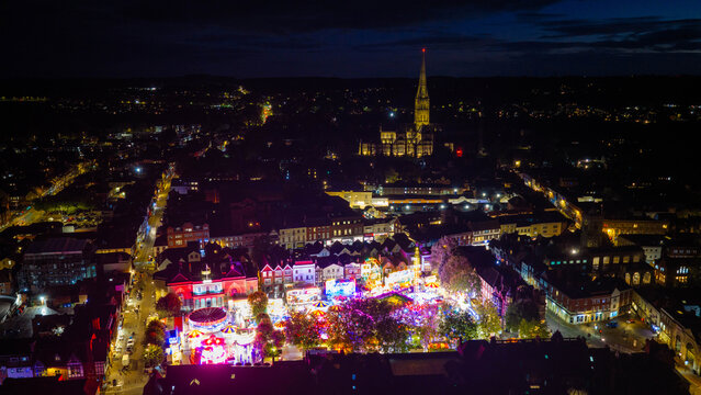Aerial view of the illuminated Salisbury Charter Fair with colorful carnival rides and stalls, set against the historic city skyline and Salisbury Cathedral in Salisbury, England, United Kingdom.