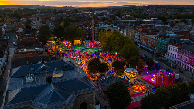 Aerial view of the Salisbury Charter Fair at dusk, featuring illuminated carnival rides, a ferris wheel, and colorful stalls set against historic architecture in Salisbury, England, United Kingdom.