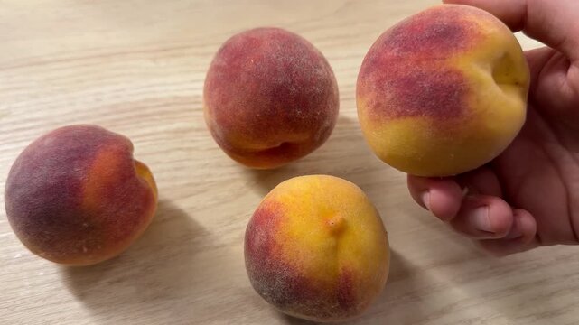 A hand lifts a red and yellow peach above three fuzzy peaches resting on a pale wood tabletop, shown in three close views