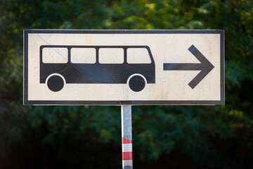 Weathered bus direction sign showing a public transportation symbol and right-pointing arrow against green vegetation. © Studio Aurelia