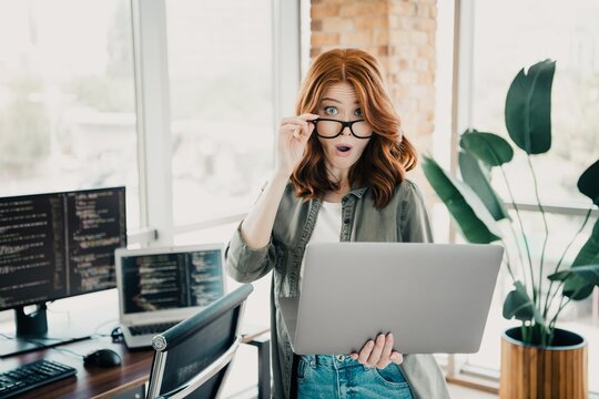 Surprised female programmer with laptop in modern office looking at screen while holding glasses
