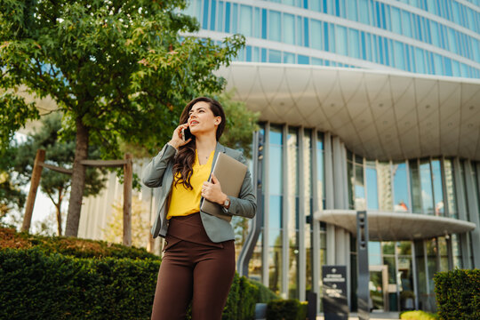 Businesswoman talking on phone carrying laptop outside office