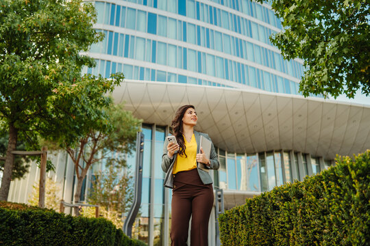 Businesswoman holding phone and laptop walking near modern office