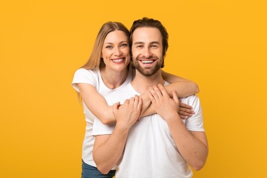 Smiling Young Caucasian Couple Embracing Each Other With Joyful Expressions In Front Of Bright Yellow Background. Captured In Indoor Studio Setting, Representing Love And Togetherness.