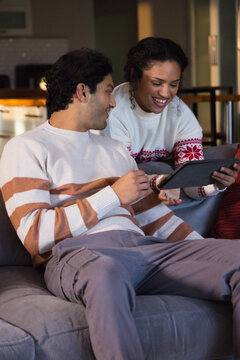 African couple sitting on gray sofa in living room, wearing white sweaters and sharing tablet