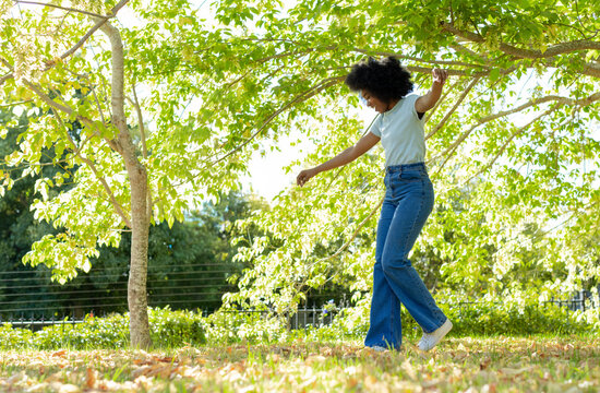 African American woman balancing on lawn in tee, jeans, sneakers while holding branch, copy space