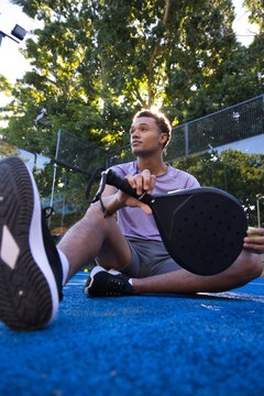 African American man sitting on blue paddle court holding black paddle, wearing purple shirt