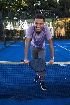 Man leaning over net on blue padel court, holding black paddle, wearing lavender tee, smartwatch
