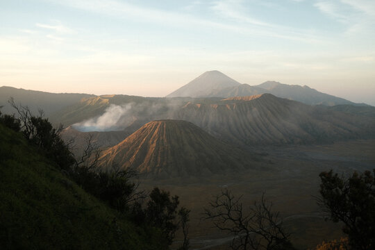 Aerial view of Mount Bromo with its volcanic crater emitting smoke, surrounded by rugged terrain and dramatic ridges under soft golden hour light in Tosari, East Java, Indonesia.