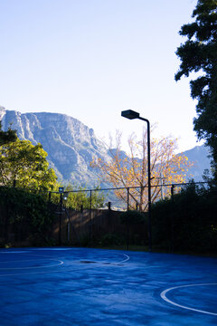 Blue sports court is reflecting low sunlight with white lines near lamp post at mountain base