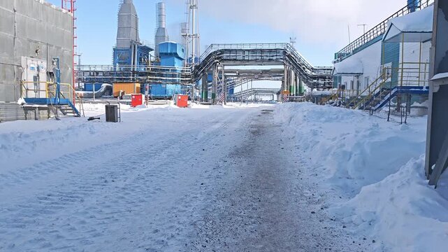Snow covered access road leads through industrial plant under pipe racks. Winter route stays clear between buildings, with road sign and metal structures framing cold site access in bright daylight.