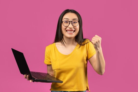 Cheerful Asian Female Student In Young Adult Age Holding Laptop With Fist Pump In Excitement At Home, Celebrating Success After Completing Study, Engaged In Online Learning Experience, Education