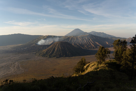 Aerial view of Mount Bromo with its smoking crater, rugged volcanic landscape, and surrounding caldera under a clear sky in Tosari, East Java, Indonesia.