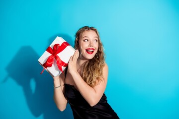 Plakat Young woman holding wrapped gift with red bow, looking surprised and happy against bright blue background