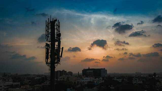 Aerial view of a network tower silhouetted against a vibrant sunset sky over an urban landscape in India.