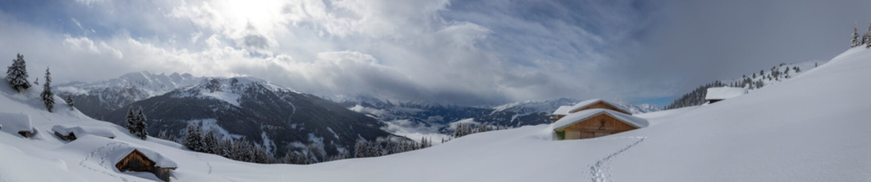 Wide panoramic view of snow-covered alpine huts in Hamberg, Zillertal, Tyrol, Austria. Winter mountain panorama with cloud inversion over valley, wooden cabins, and snowy peaks in Austrian Alps.