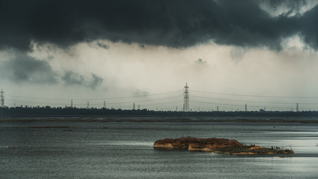 Aerial view of a small island in a vast lake under a dramatic sky filled with dark storm clouds and power lines stretching across the horizon in India.