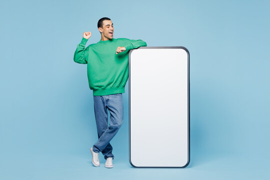Full body young middle eastern man wear green shirt casual clothes big huge blank screen mobile cell phone smartphone with copy space mockup area do winner gesture isolated on plain blue background