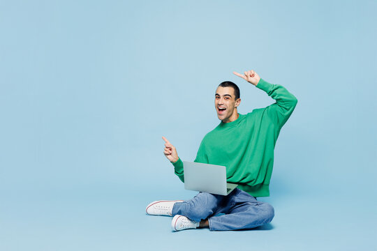 Full body young IT man he wear shirt white t-shirt casual clothes sit hold use work on laptop pc computer point finger aside isolated on plain blue cyan background studio portrait. Lifestyle concept.