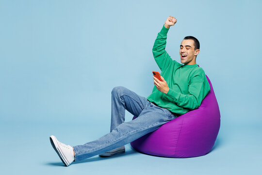 Full body young middle eastern man wear green shirt casual clothes sit bag chair hold in hand use mobile cell phone do winner gesture isolated on plain light blue cyan background. Lifestyle concept