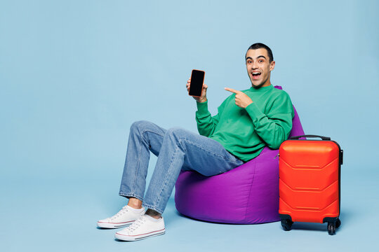 Traveler man wear casual clothes sit bag chair near suitcase use blank screen mobile cell phone isolated on plain blue background. Tourist travel abroad in free time getaway Air flight trip concept