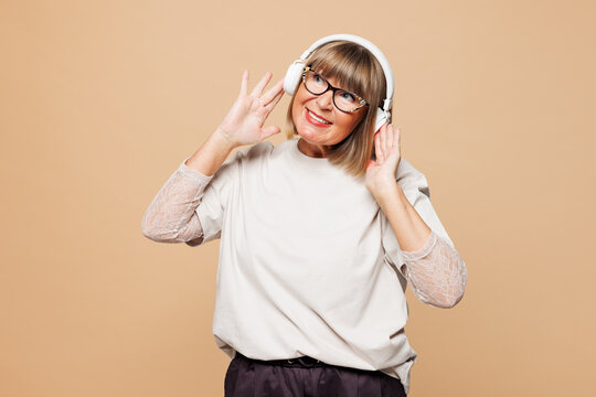 Elderly fun blonde woman 50s years old she wear white shirt casual clothes listen to music in headphones look aside isolated on plain pastel light beige background studio portrait. Lifestyle concept.