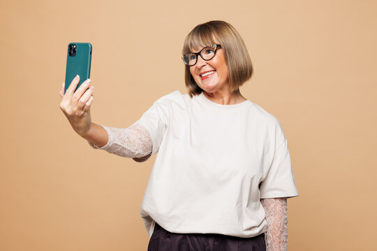 Elderly blonde woman 50s years old wear white shirt casual clothes do selfie shot on mobile cell phone post photo on social network isolated on plain pastel light beige background. Lifestyle concept.