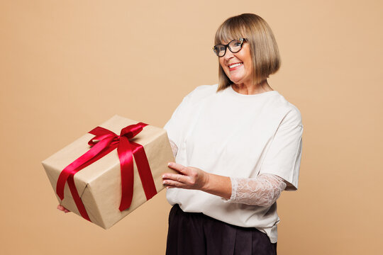 Elderly happy blonde woman 50s years old wear white shirt casual clothes hold give present box with gift ribbon bow isolated on plain pastel light beige background studio portrait. Lifestyle concept.