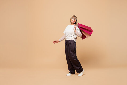 Full body elderly happy young woman wear casual clothes hold in hand paper package bags after shopping look aside isolated on plain pastel light beige background. Black Friday sale buy day concept