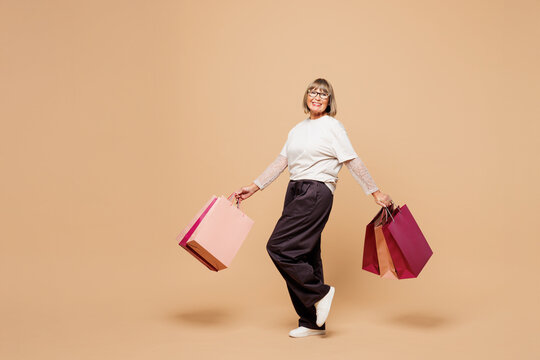 Full body elderly happy fun young woman wears casual clothes hold in hand paper package bags after shopping walk go isolated on plain pastel light beige background. Black Friday sale buy day concept