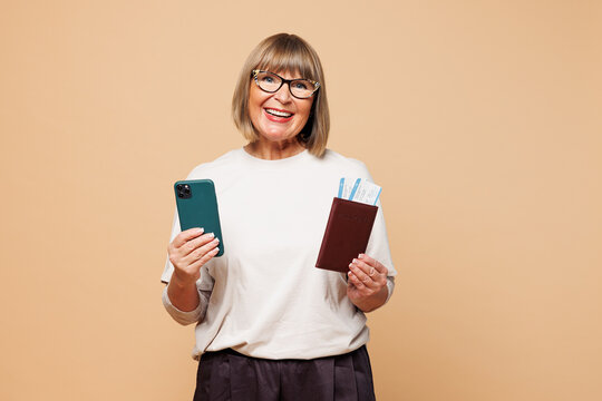 Traveler elderly woman in casual clothes hold passport ticket use mobile cell phone isolated on plain beige background. Tourist travel abroad in free time rest getaway Air flight trip journey concept