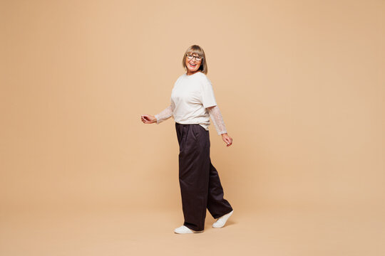 Full body side view smiling happy elderly blonde woman 50s years old she wear white shirt casual clothes walk go look camera isolated on plain pastel light beige background studio. Lifestyle concept.