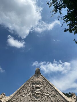 Carved stone gable of a traditional Javanese building with Kala face relief and floral ornament, seen from below against a blue sky with white clouds and tree branches