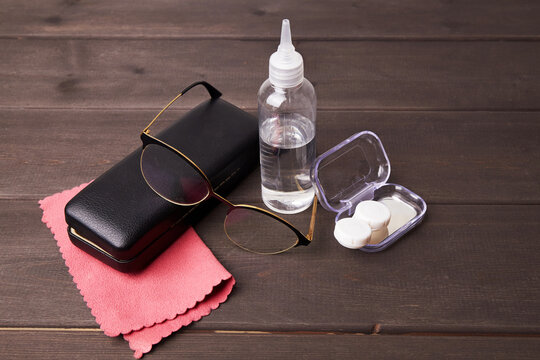 Eyeglasses sit next to a cleaning cloth and a bottle of lens solution on a wooden table.