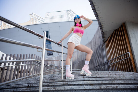 Young woman posing on pink roller skates on urban steps wearing sporty summer outfit, headphones and cap for confident street style and leisure skating