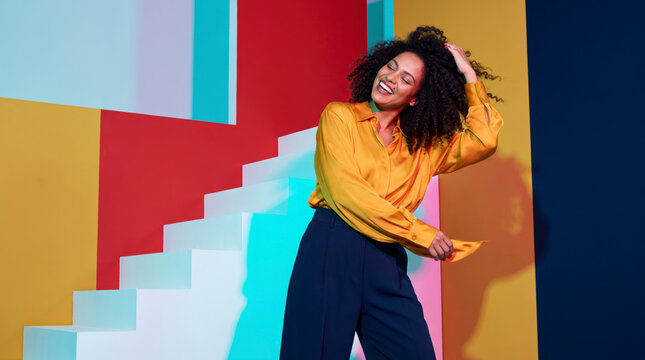 Uninhibited woman with curly hair laughing in saffron blouse beside white staircase and red wall
