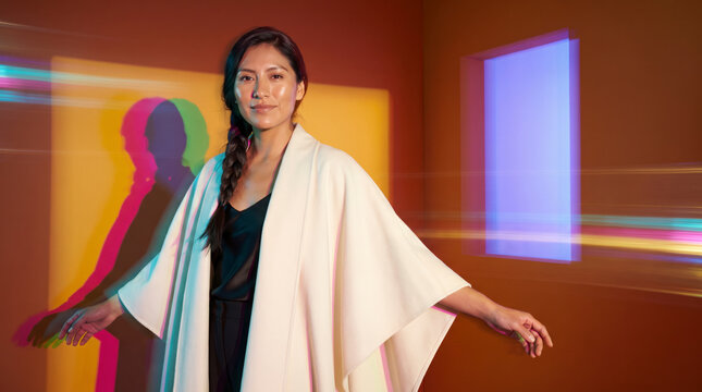 Composed woman with long braid in ivory cape coat among colorful light trails in orange room