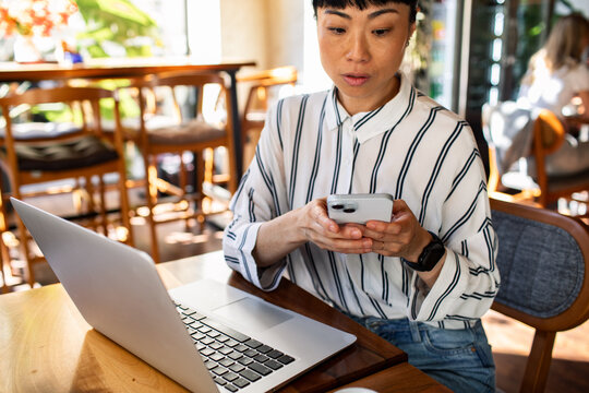 Woman texting on smartphone while working on laptop in cafe