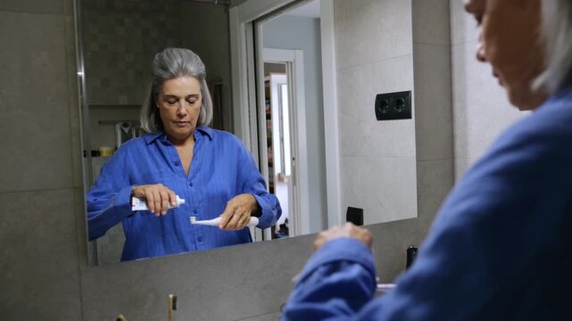 Elderly woman with grey hair putting toothpaste on her electric toothbrush and brushing her teeth