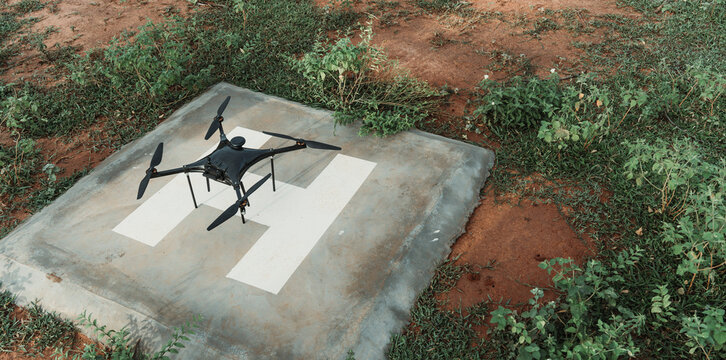View of a black drone positioned on a concrete helipad with white markings, surrounded by patches of green grass and dry earth in India.